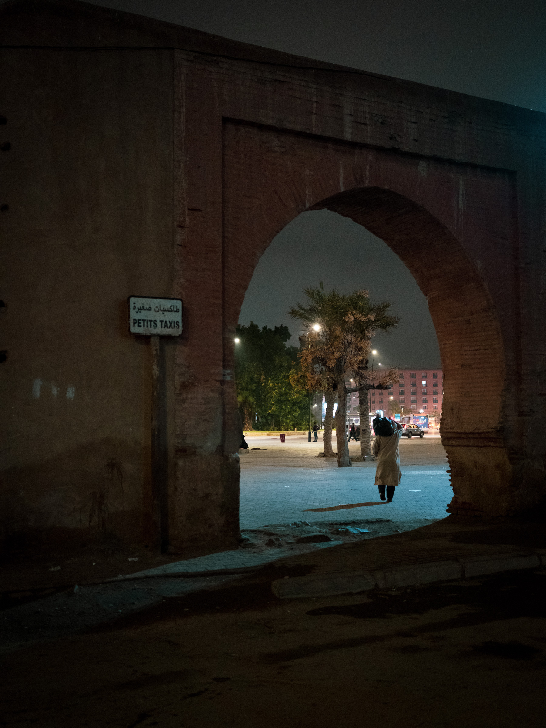 A figure passes through one of the old city gates at night, a "Petits Taxis" sign fixed to the ancient brick arch. Beyond: trees, streetlights, and an empty boulevard.