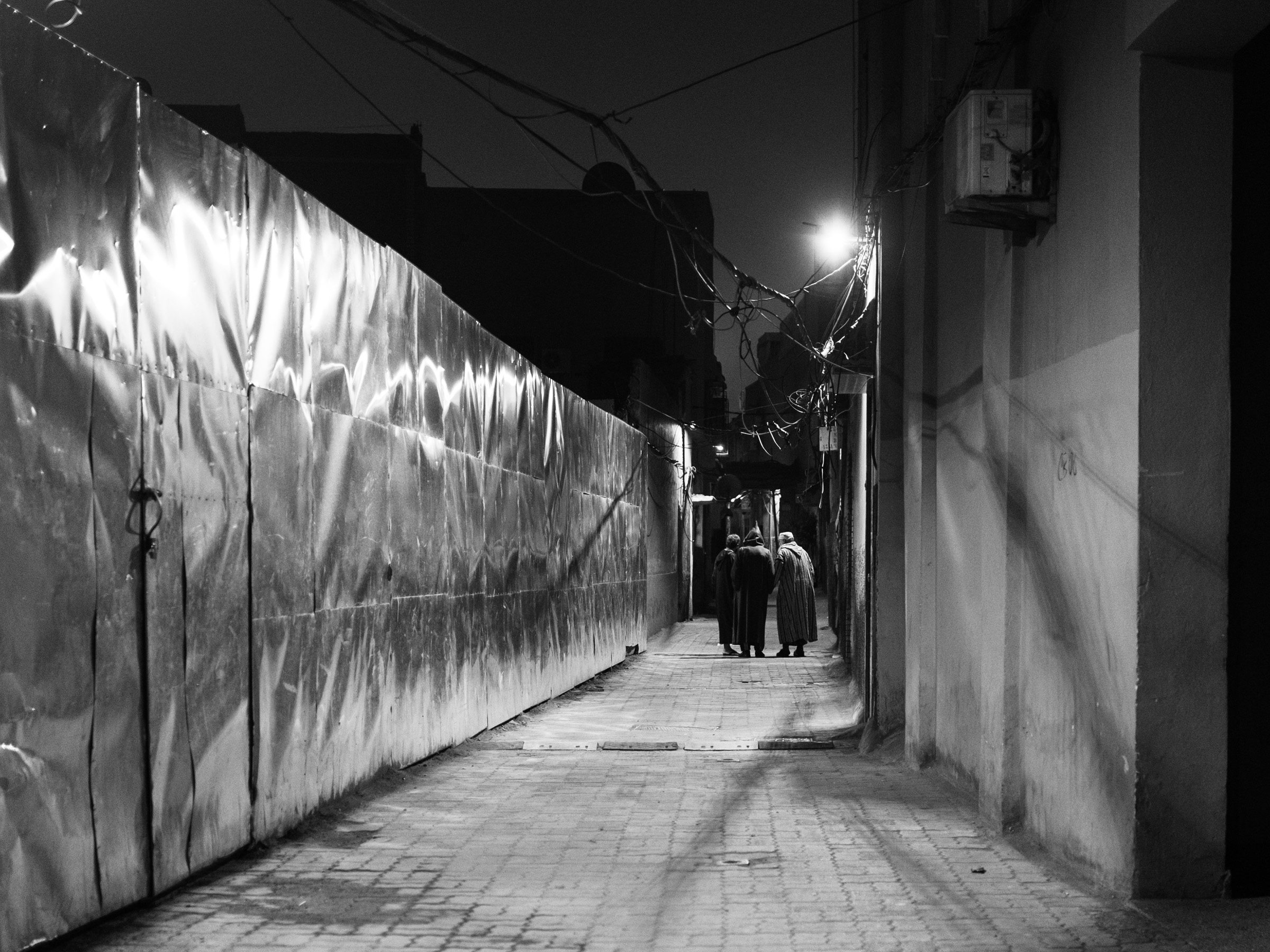 Three men in djellabas walk together through a narrow lane, a construction hoarding catching the streetlight on the left. Likely heading to morning prayer.