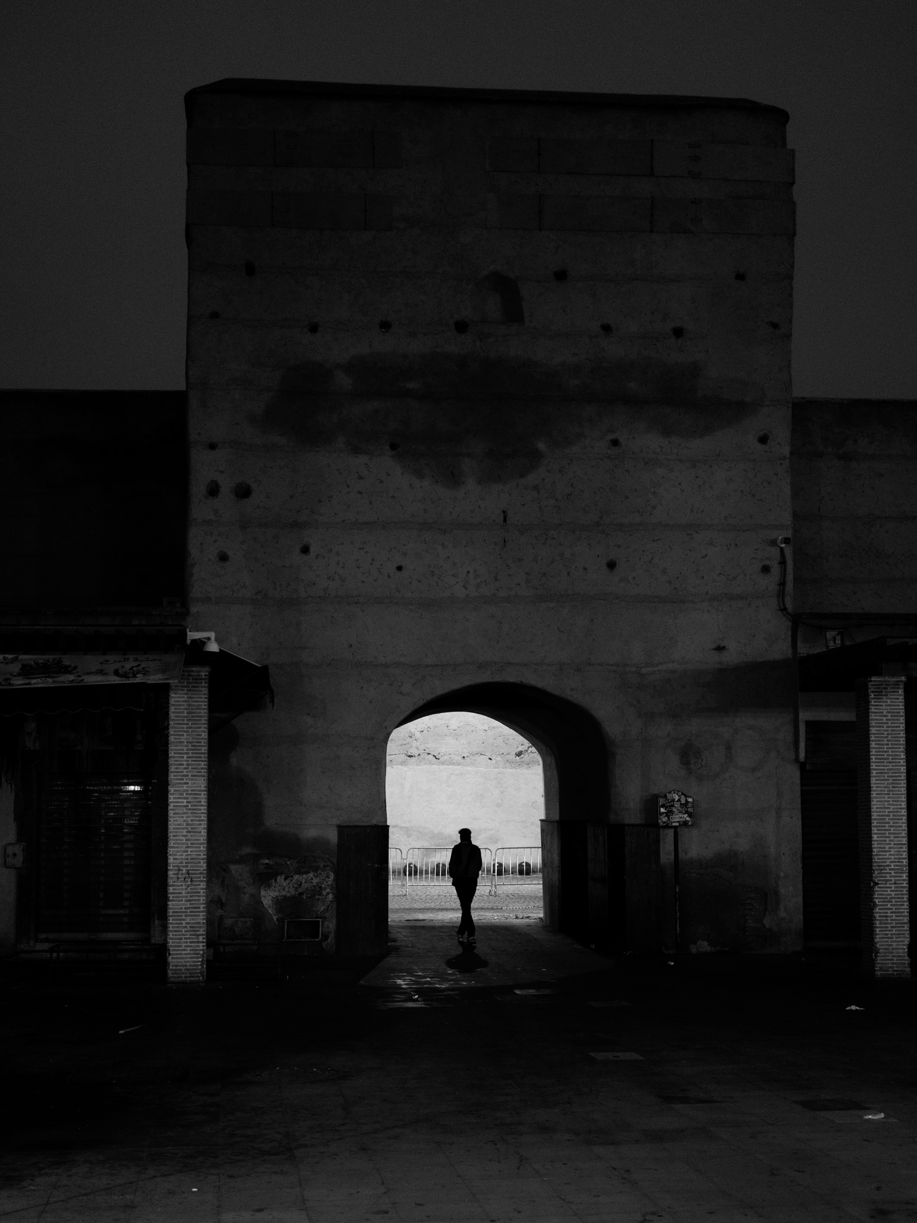 A lone figure stands framed in the arch of a massive city gate, backlit against the pre-dawn light beyond. The dark, weathered stone amplifies the sense of threshold and solitude.