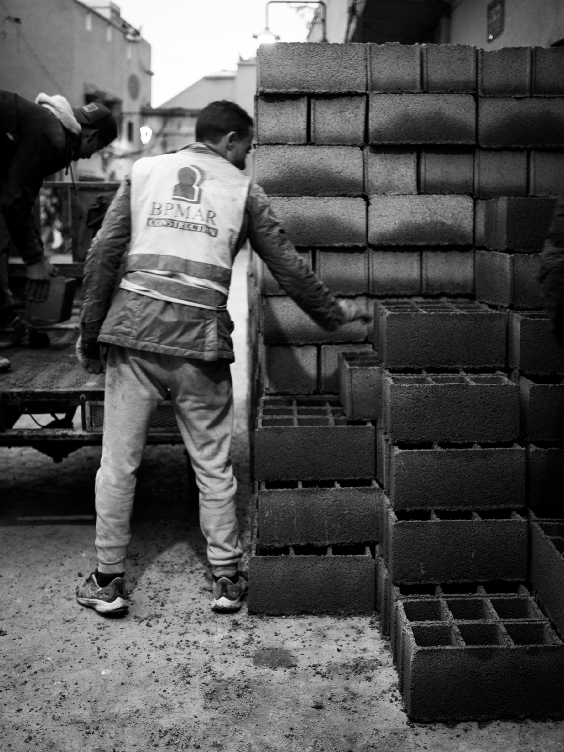 A construction worker in a hi-vis vest unloads concrete blocks from a truck in a narrow Medina street. Friday morning, but work continues.