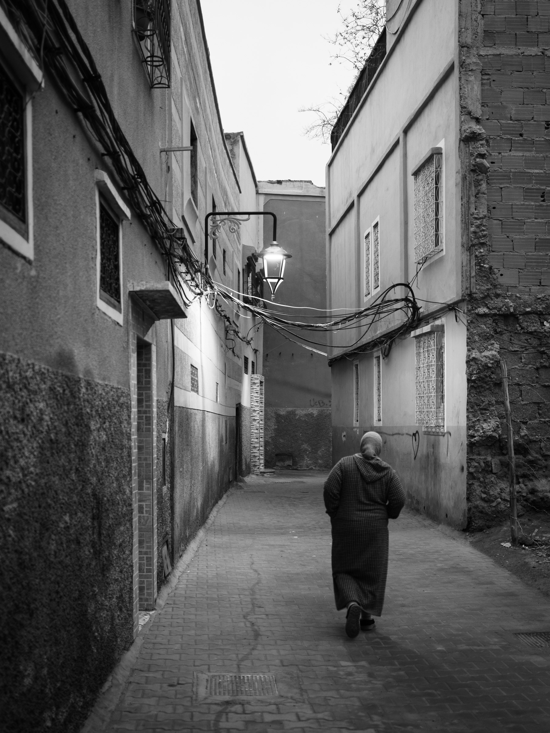 A woman in a striped djellaba walks away through a narrow Medina alley, a still-lit lantern above her. Dawn has arrived but the streetlight hasn't noticed yet.