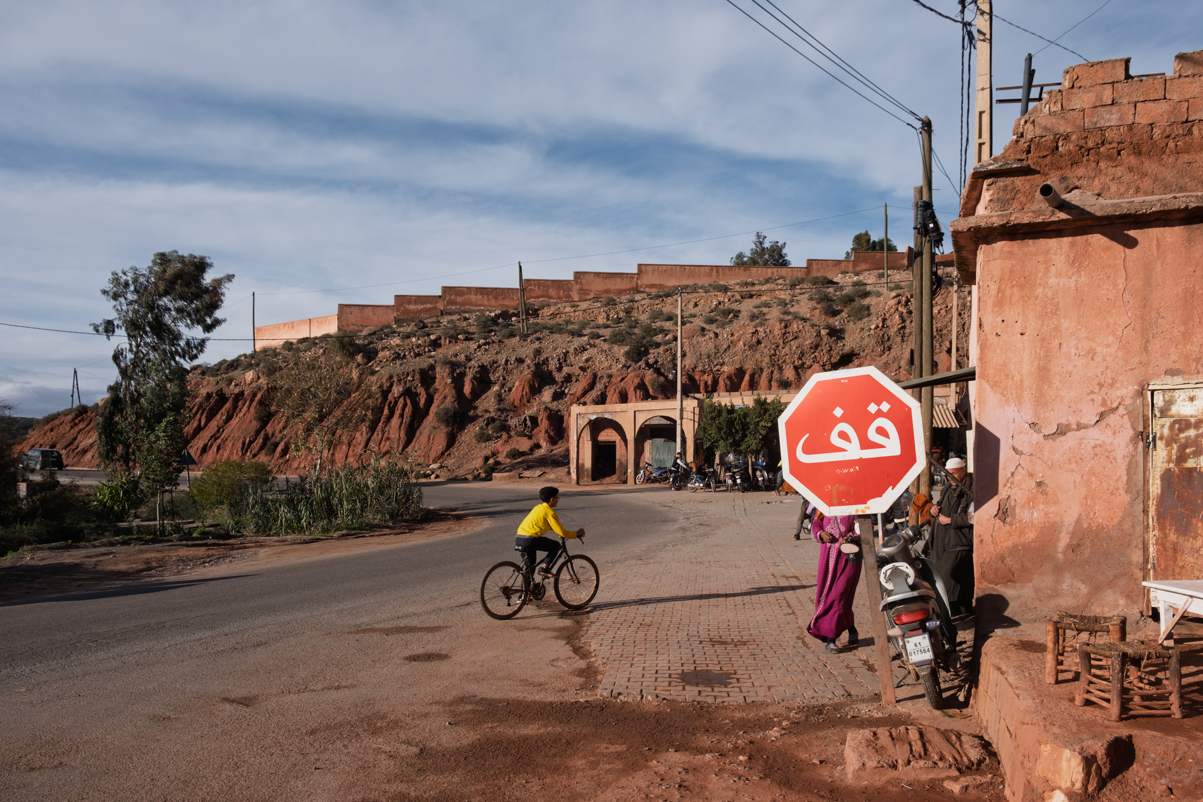 Child riding a bicycle on a street in a Moroccan village, with red clay hills in the background.