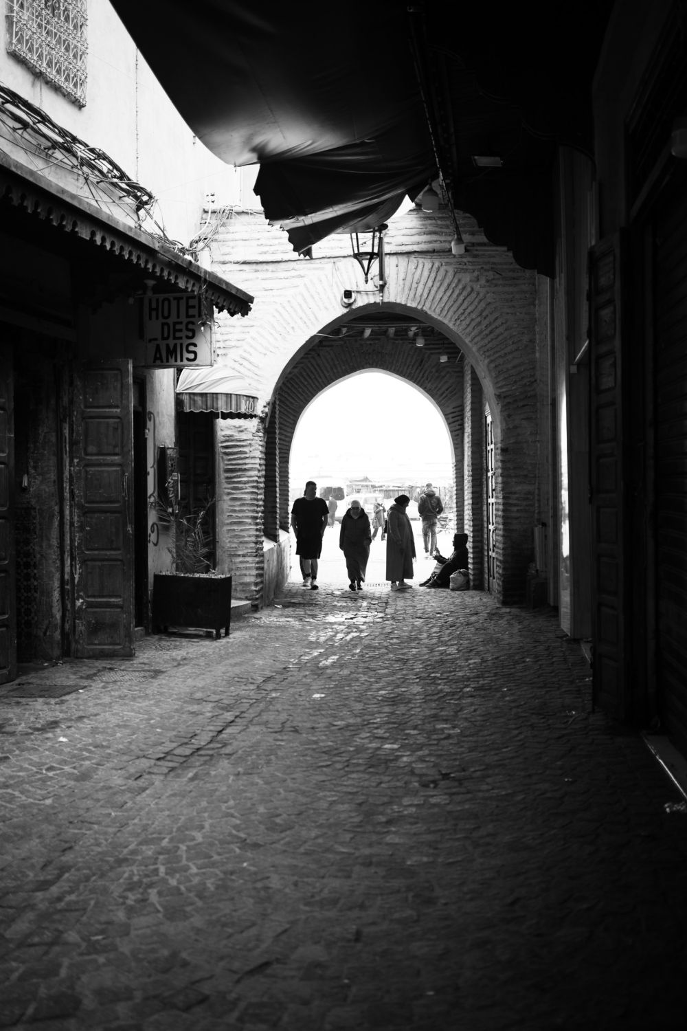 Several figures walk through a brick arch leading onto Djemaa el-Fna, backlit by the open sky beyond. The sign for Hôtel des Amis is visible on the left wall.