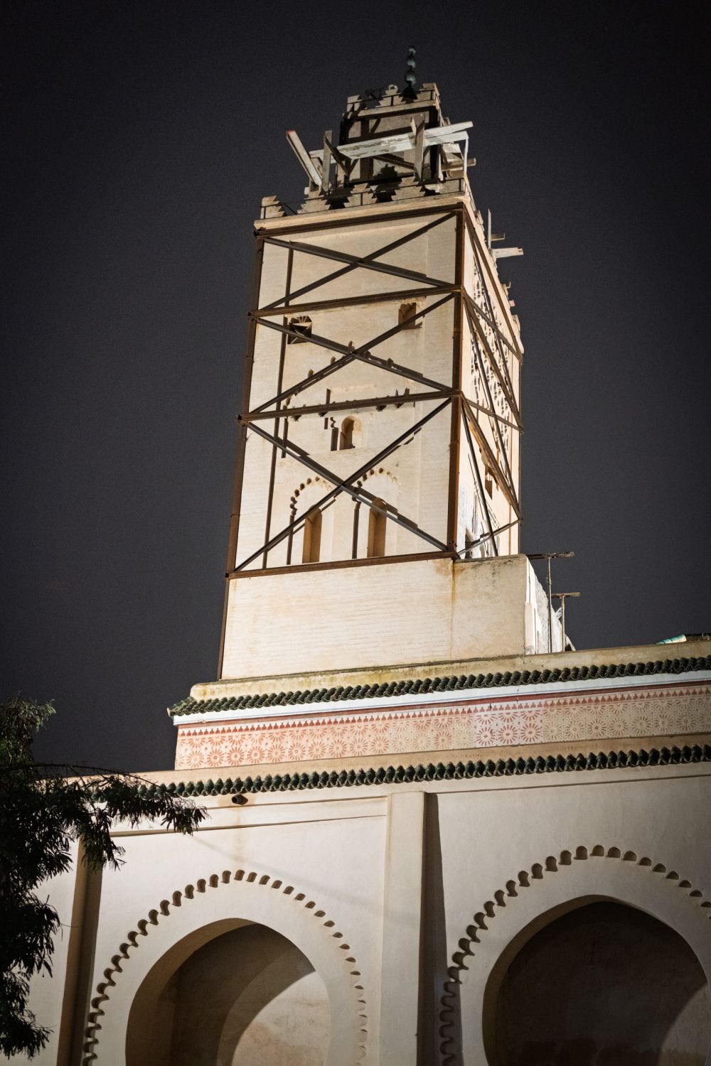 A mosque minaret wrapped in restoration scaffolding, photographed against the night sky. The geometric ornamental band at its base remains intact.