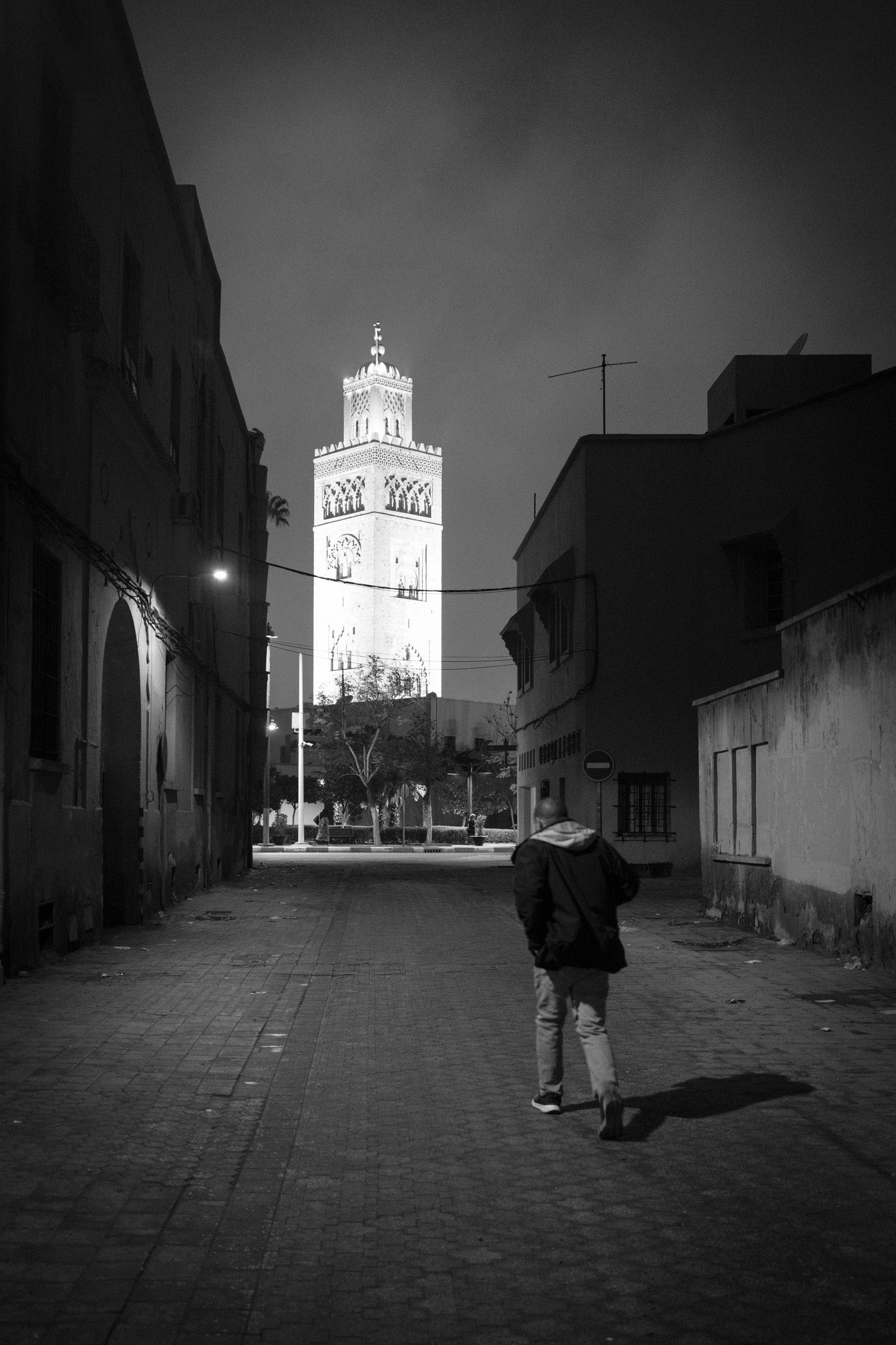 A lone figure walks through a deserted alley in the Medina, the illuminated minaret of the Koutoubia Mosque rising at the end of the street. Early Friday morning, Marrakech.