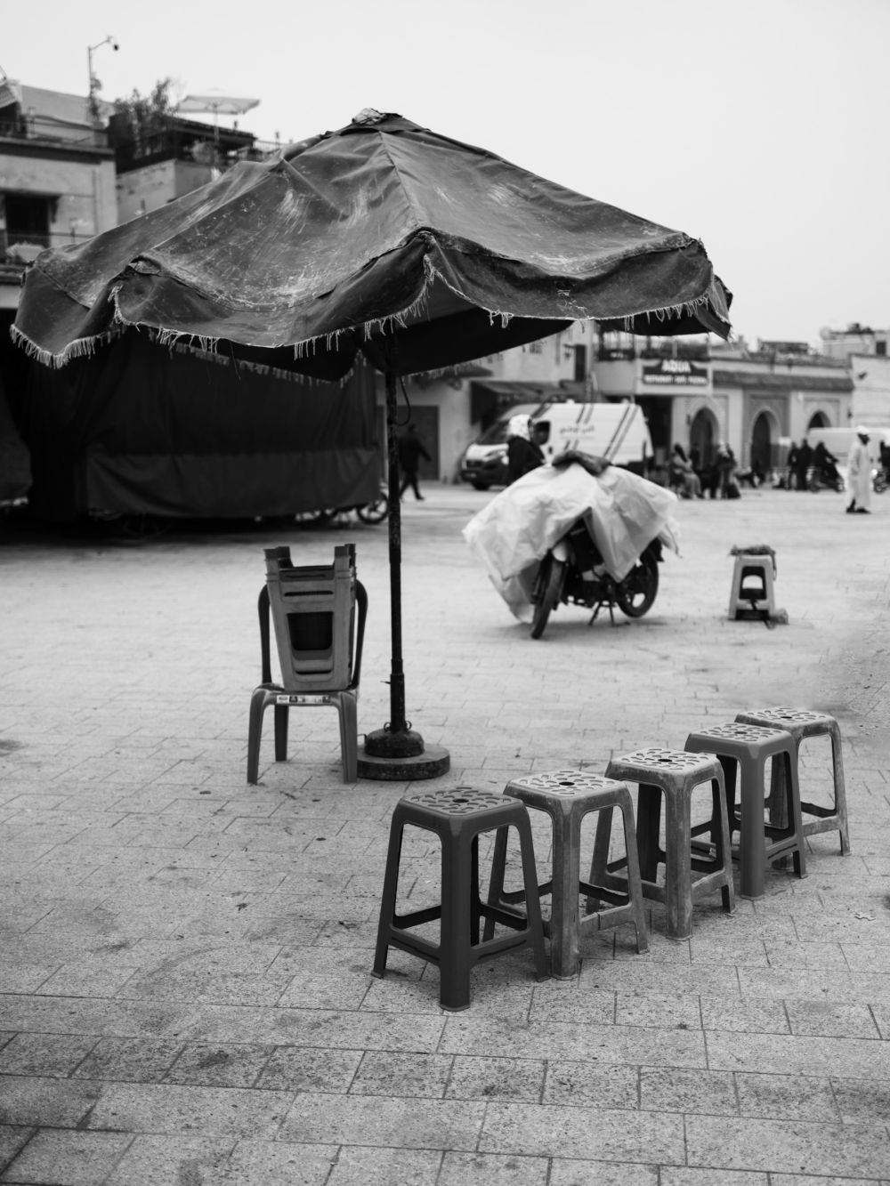 Plastic stools arranged around a worn market umbrella on Djemaa el-Fna. The square is slowly waking — a motorbike covered in plastic sheeting stands in the background.