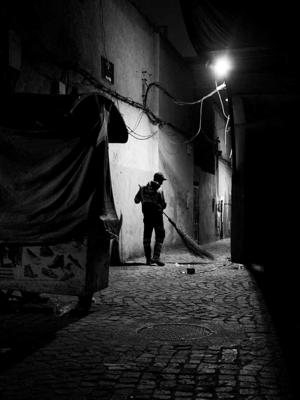 A worker sweeps a narrow alley in the Medina, lit from above by a single bare bulb. Closed market stalls and tangled overhead cables line the passage.