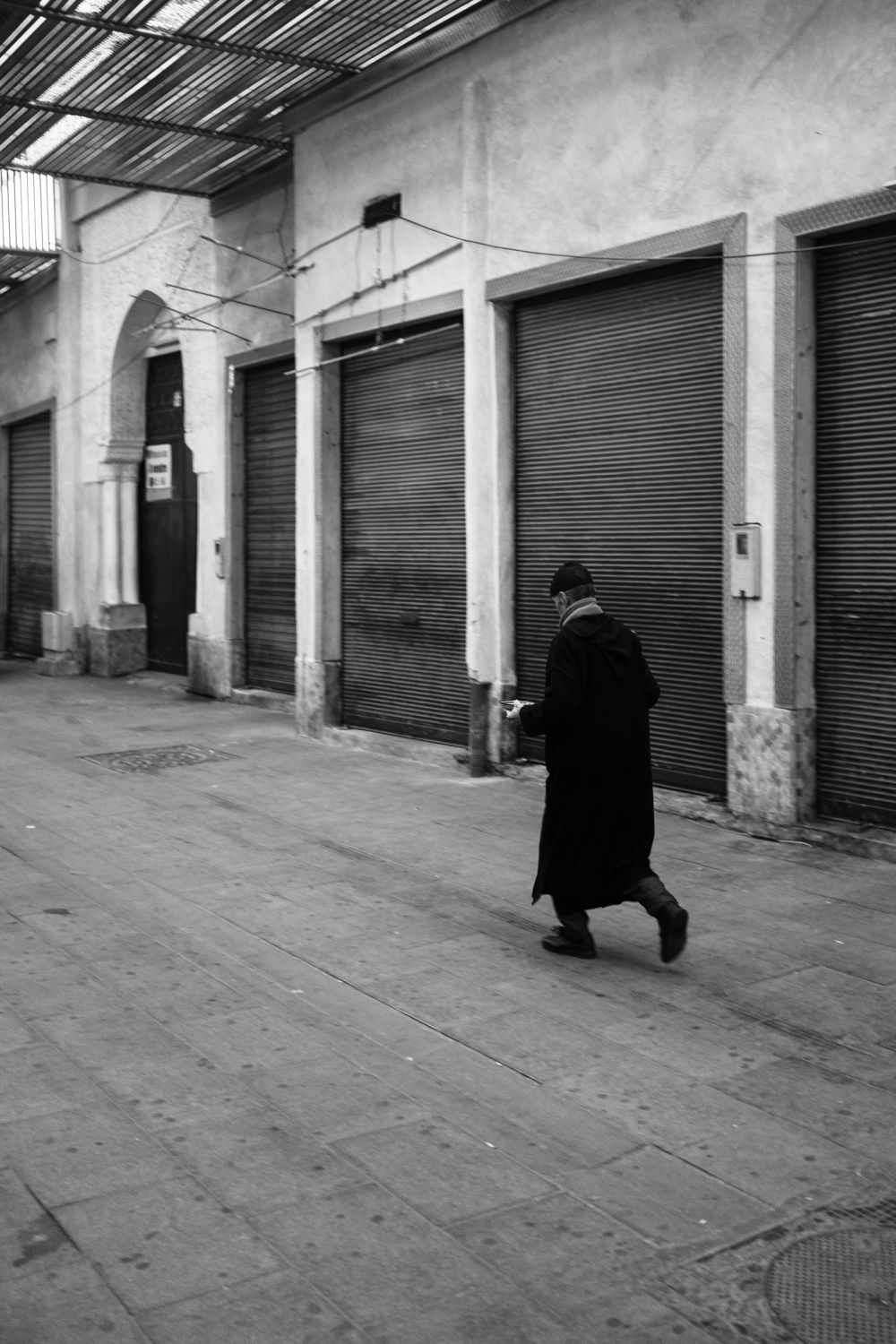 A man in a long dark djellaba walks past a row of shuttered stalls under a corrugated canopy. The market is closed, the passage almost empty.