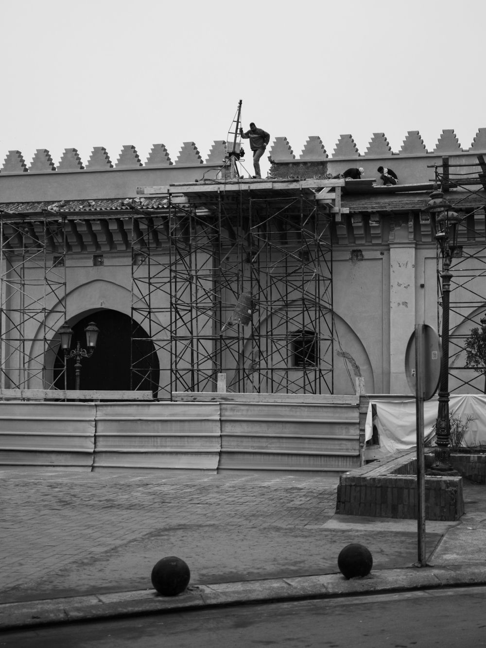 Workers on the roof of the same building, one operating a small crane or winch. A closer view of the scaffolding and the merloned parapet above.