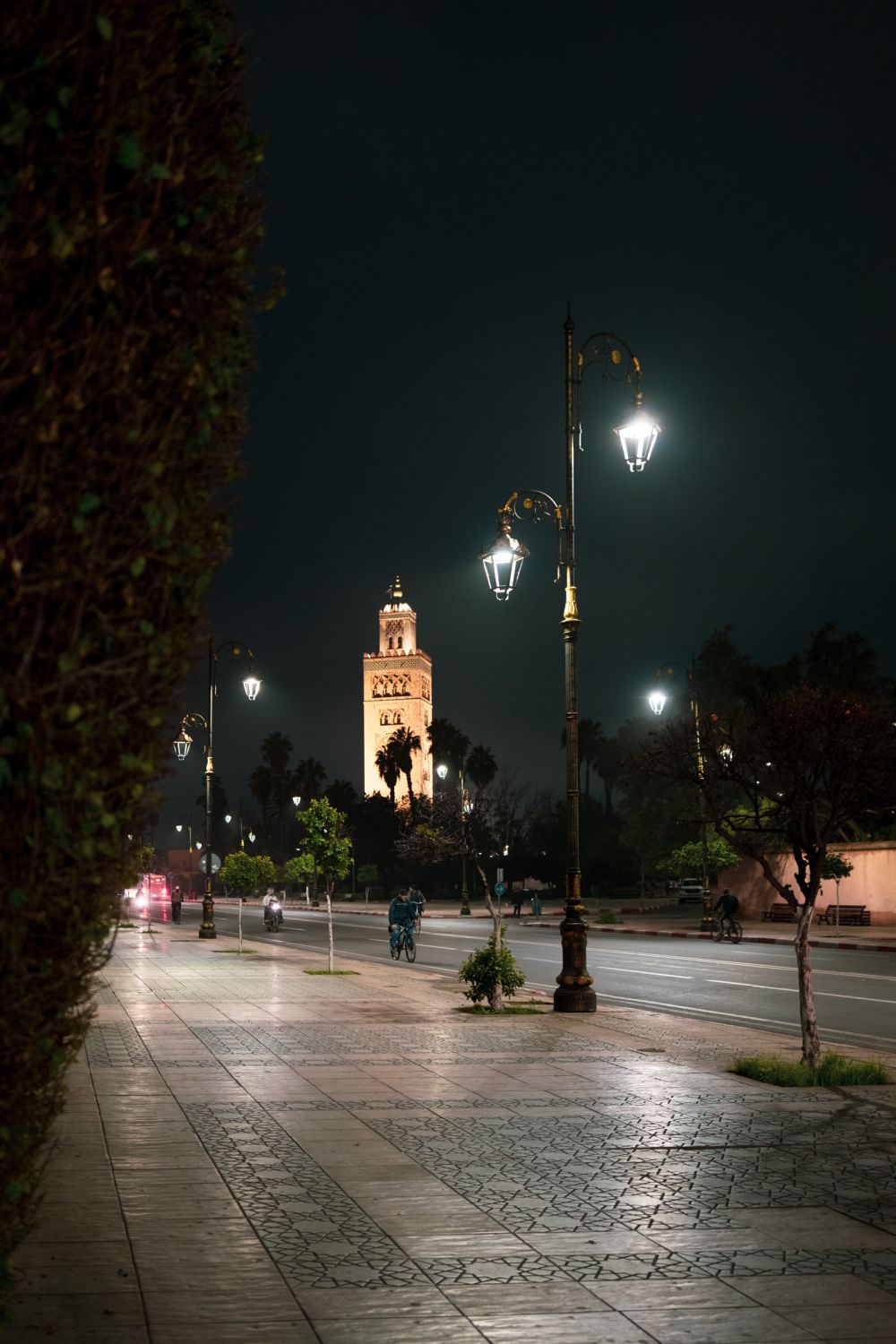 The Koutoubia Mosque minaret glows in the distance beyond an empty boulevard, ornate street lamps lining the promenade. Cyclists pass below, the city barely stirring.