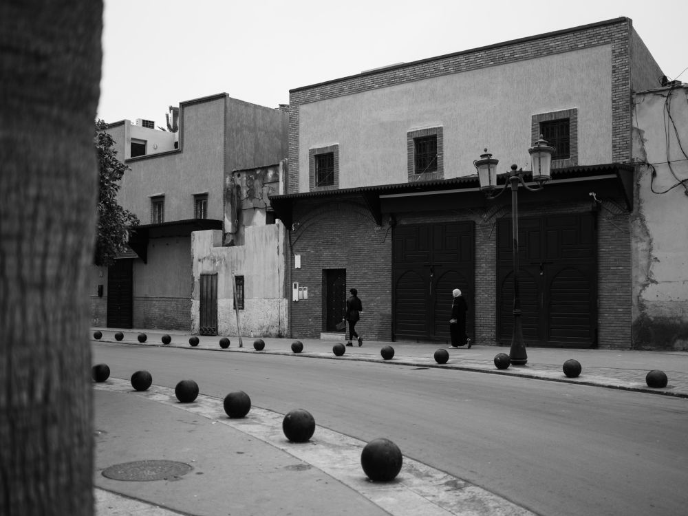A row of stone bollards lines a quiet street near the square, a woman on a scooter and another on foot passing a row of shuttered buildings behind.
