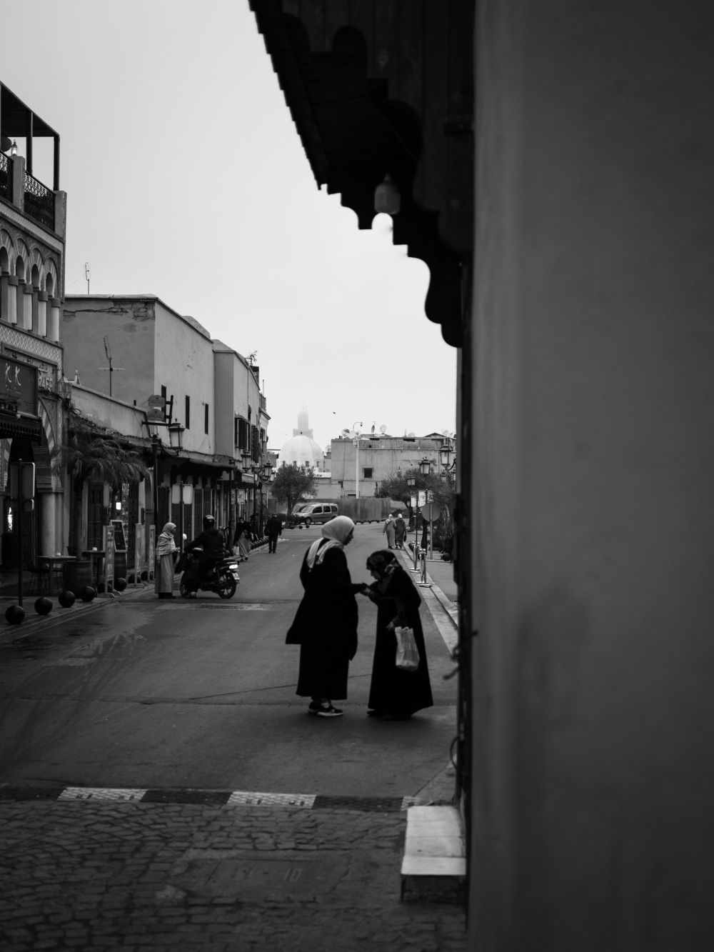 Two women stop to talk on a quiet street near Djemaa el-Fna, framed by a Moorish doorway arch. A mosque dome floats in the haze in the distance.