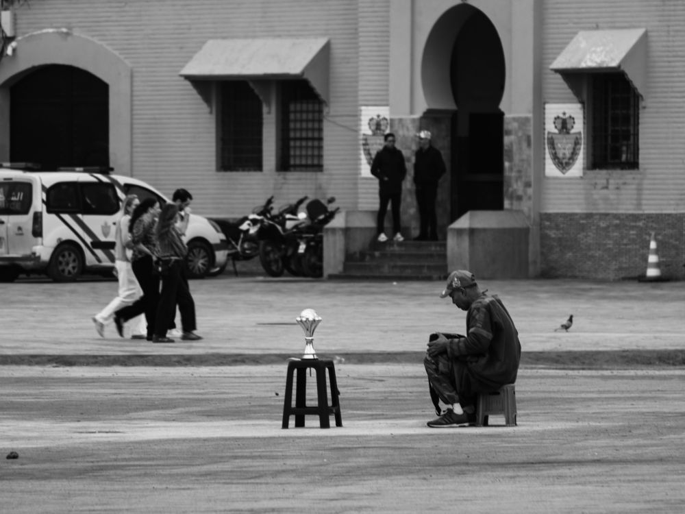 A man crouches alone on the square beside a small trophy on a stool, apparently preparing his act. In the background, groups of people pass in front of a public building.